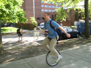 Daniel riding a unicycle along a campus sidewalk, wearing a backpack.